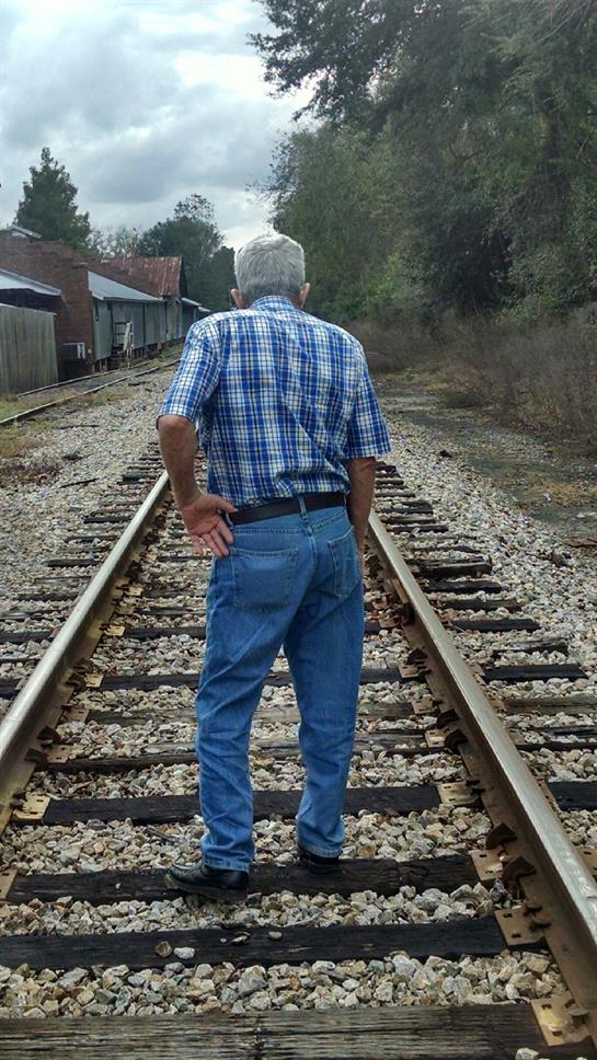 An elderly man in a plaid shirt stands on railway tracks, reflecting quietly.