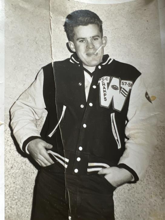 A young man in a vintage jacket stands confidently in a 1950s community space.