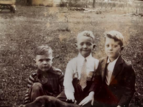 Three boys sit together on the grass with a dog; they wear vintage clothing from the 1940s.