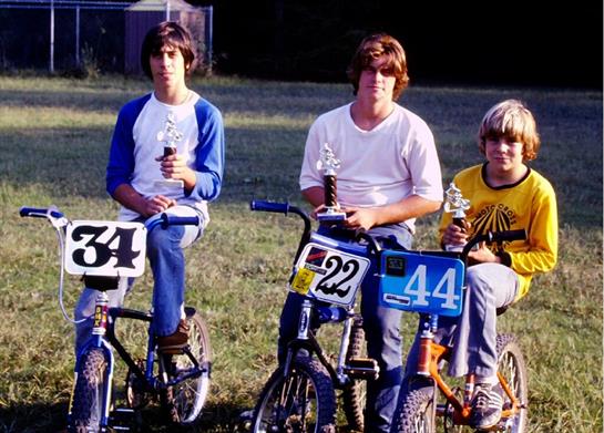 Three boys sit on their BMX bikes holding trophies after winning a race in the summer sunlight.