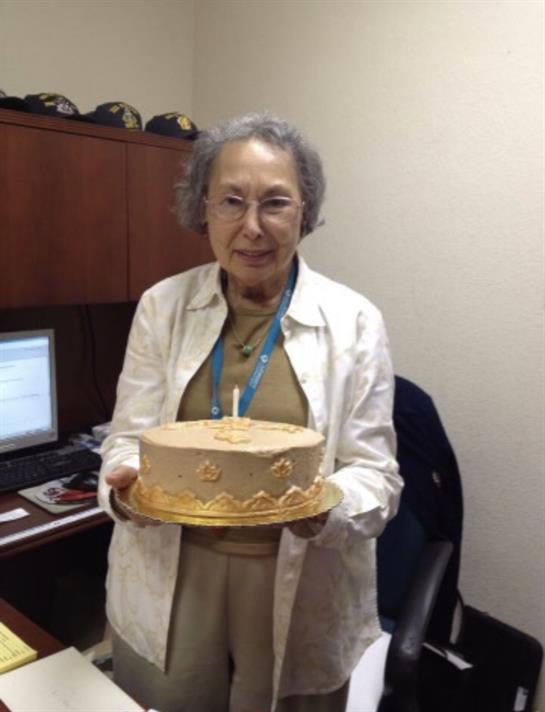 A woman smiles while holding a beautifully decorated cake in an office environment.