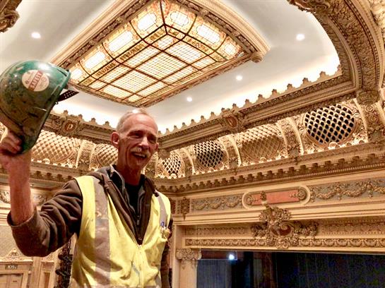 A worker smiles while holding tools during restoration of a richly detailed theater interior.