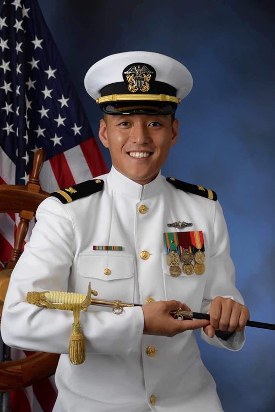 Young naval officer smiles while holding a ceremonial sword, dressed in pristine white uniform.