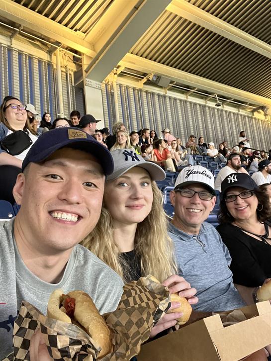 Group of friends at a baseball game share food and laughter in a lively atmosphere.