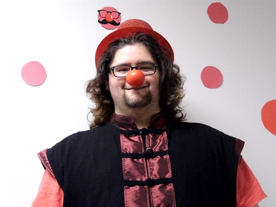 A clown wearing a red nose and hat smiles brightly in a decorated room with pink polka dots.