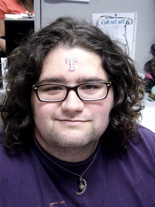 A young man with curly hair and glasses shows off his forehead tattoo at a casual gathering.