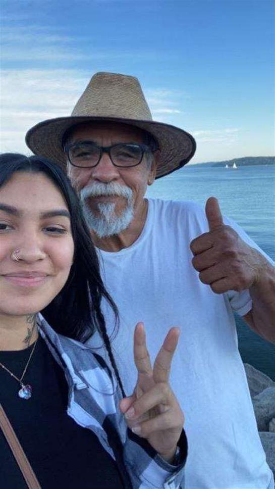 A young woman poses playfully with an elderly man against a serene waterfront backdrop at sunset.