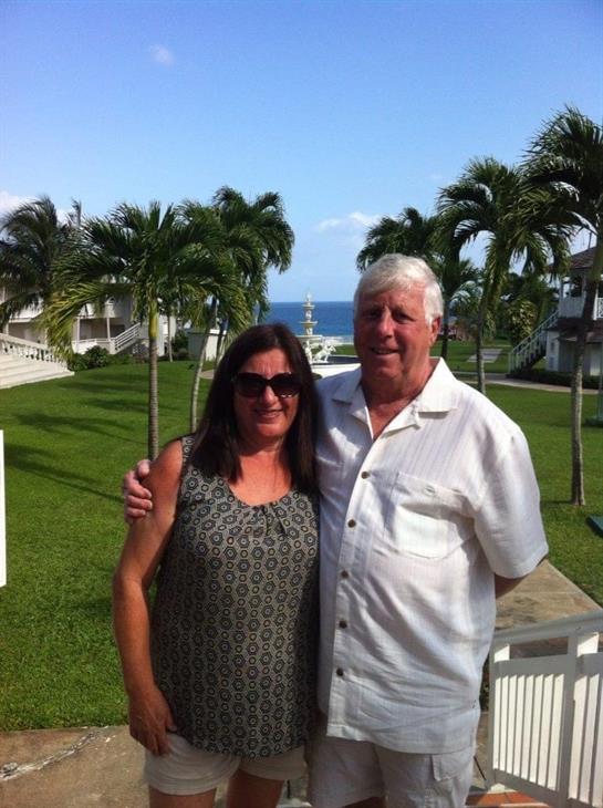 A couple stands together at a tropical resort, smiling against a beautiful ocean backdrop.