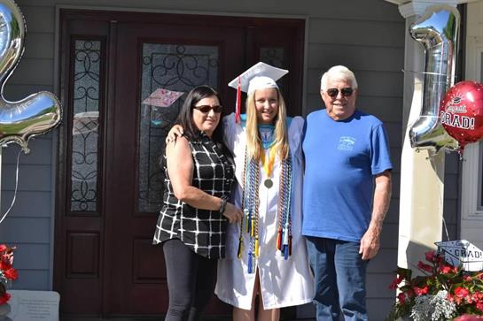 Young woman dressed in graduation attire poses with parents in front of their home.