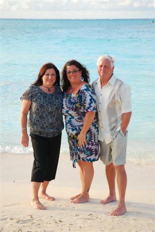 Three adults stand on a sandy beach with clear blue water, smiling and enjoying a sunny day.
