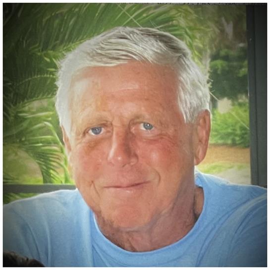 Elderly man with silver hair and blue eyes smiles warmly while sitting in a lush garden.