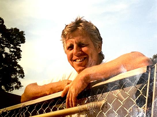 A man with short hair smiles while leaning on a chain-link fence under clear skies.