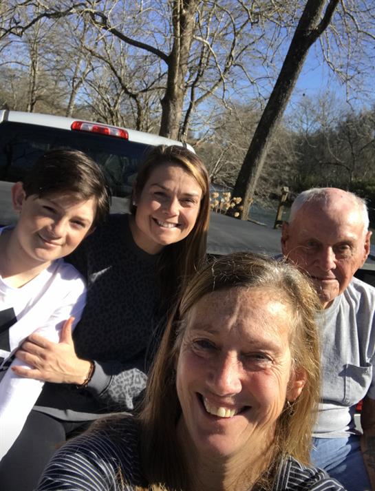 Four family members gather for a cheerful group selfie on a bright, sunny day surrounded by nature.