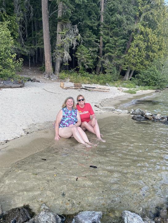 Two friends sit in shallow water by the shore, enjoying a sunny day surrounded by trees.