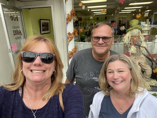 Three friends smile happily outside a shop decorated with autumn accents and seasonal decor.