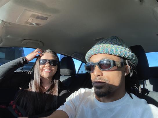 A couple smiles and poses for a selfie while driving in bright sunlight, showcasing their joy.