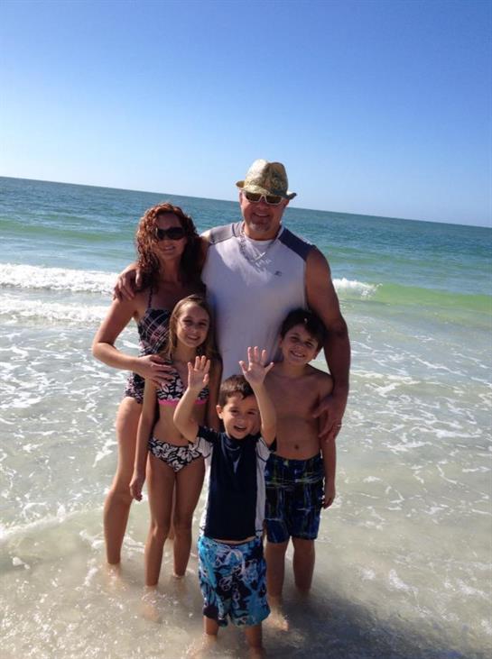 A family stands together on the beach, waving at the camera with ocean waves in the background.