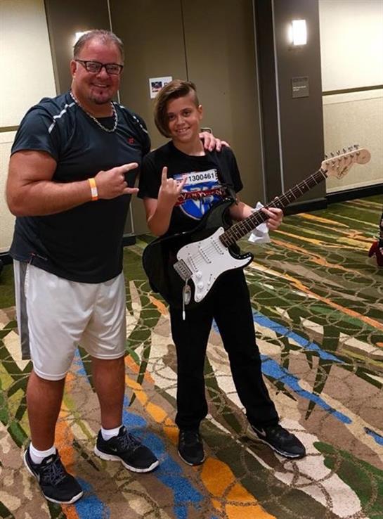 A young guitarist stands proudly with an adult, both making rock signs in a hotel lobby.