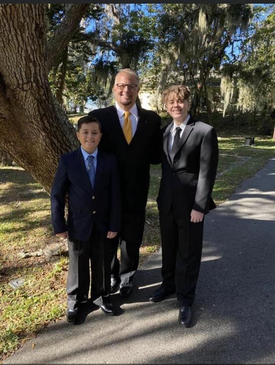 Three men in suits pose together outside in a park, enjoying a bright, sunny day.