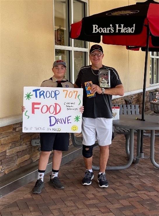 Two individuals stand with a sign advertising a food drive to support their troop.