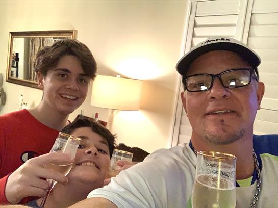 Two young boys and an adult smile while holding sparkling drinks at a cozy home gathering.