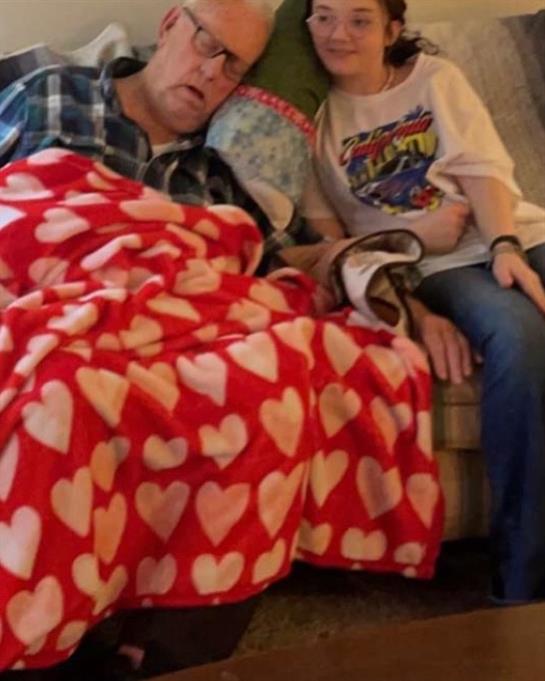 Two family members sit closely on a couch, wrapped in a blanket, enjoying a warm, intimate moment.
