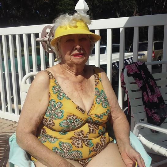 An elderly woman sits on a chair by the pool, enjoying the sunshine in a vibrant swimsuit.
