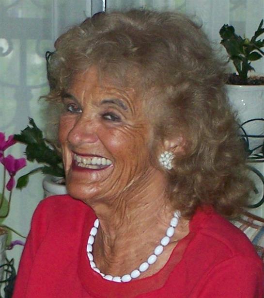 Elderly woman with curly hair smiles warmly while surrounded by indoor plants and flowers.