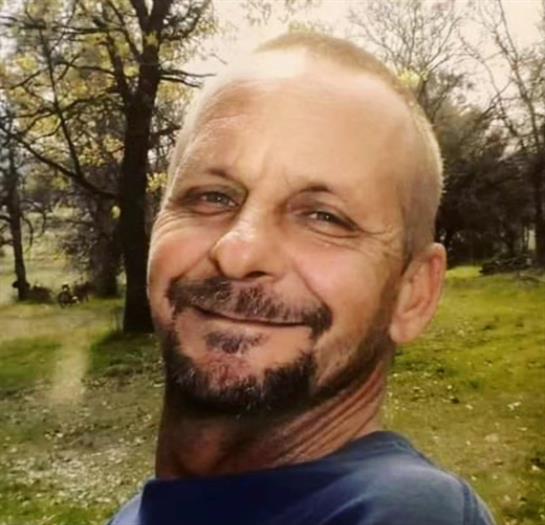 A man smiles brightly while standing in a park surrounded by trees and grass, enjoying the day.