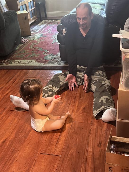 Child plays with a toy while sitting on the floor, interacting with a seated adult in a warm home.