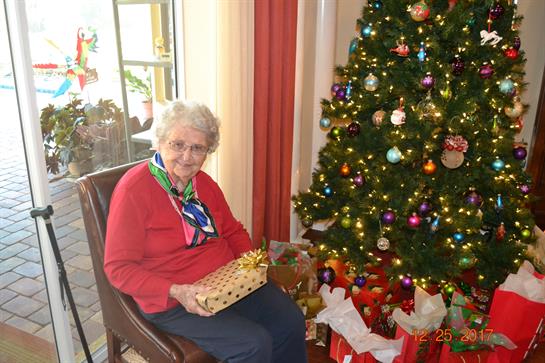 An elderly woman beams with a gift by a decorated Christmas tree and presents.
