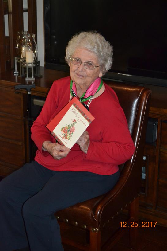 Senior woman warmly smiles while holding a colorful greeting card, seated in a wooden chair.