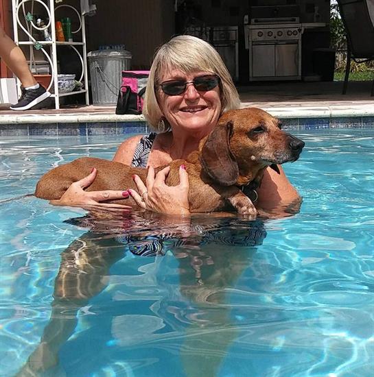 A smiling woman holds a brown dog while floating in a clear pool on a sunny day.