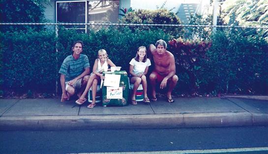 Group of two adults and two children relax by a lemonade stand on a warm day in a neighborhood.