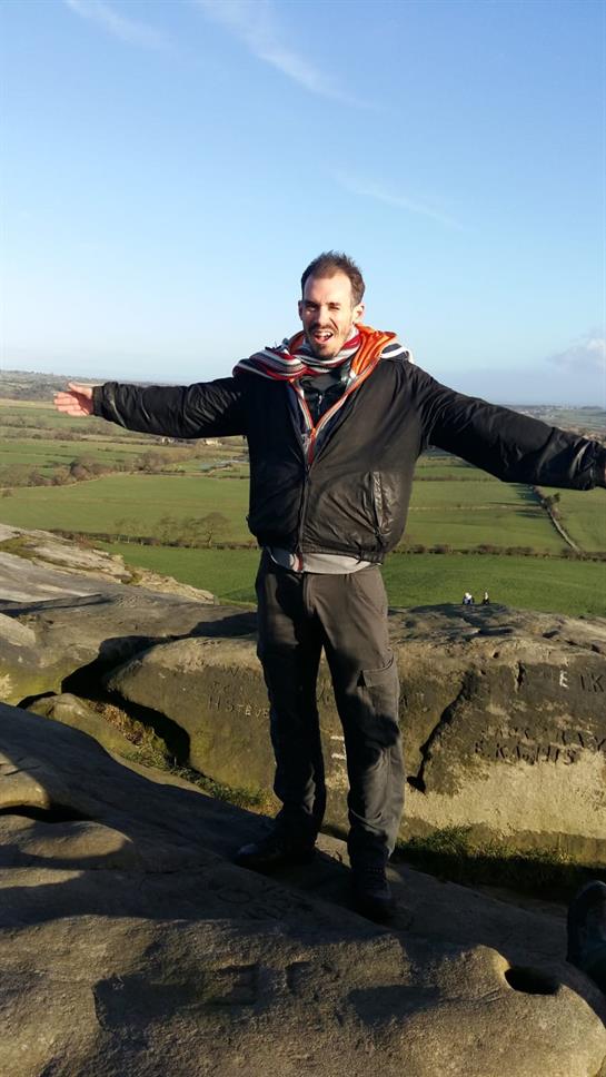 A man stands on a rocky ledge with arms wide open, admiring the vast green landscape below.