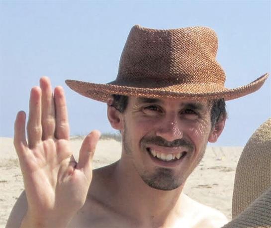A cheerful man in a straw hat waves at the camera while enjoying a sunny day at the beach.