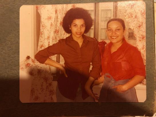 Two women stand confidently in a warmly lit room, showcasing their vibrant outfits and friendship.