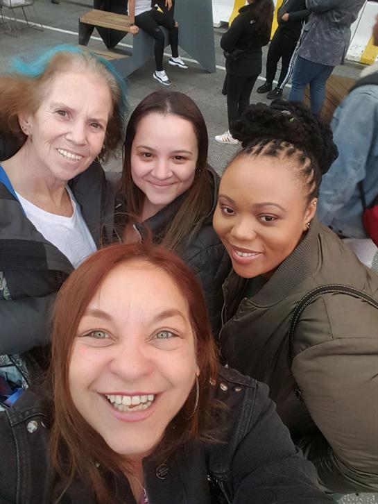 Four friends smile happily while taking a selfie in a lively outdoor setting with crowds.