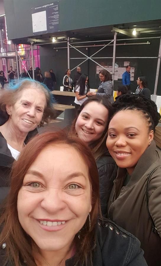Four women are smiling and taking a cheerful selfie while surrounded by friends at an event.