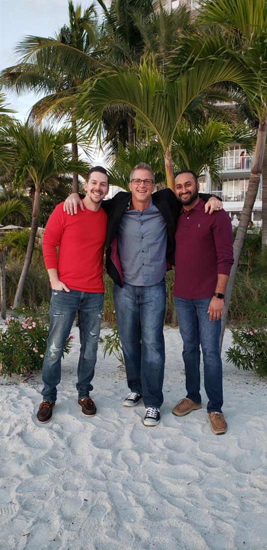 Three men stand together on a beach, smiling and enjoying their time during a sunny day.
