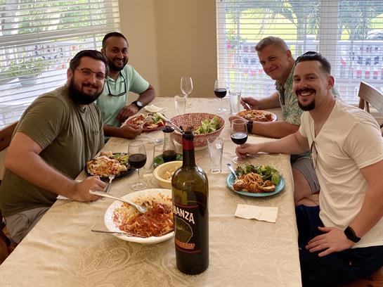Four friends share a meal at a dining table filled with pasta, salad, and wine glasses.