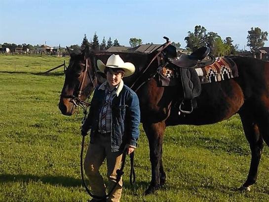 A cowboy in a hat poses with his horse in a lush green field under a clear blue sky.