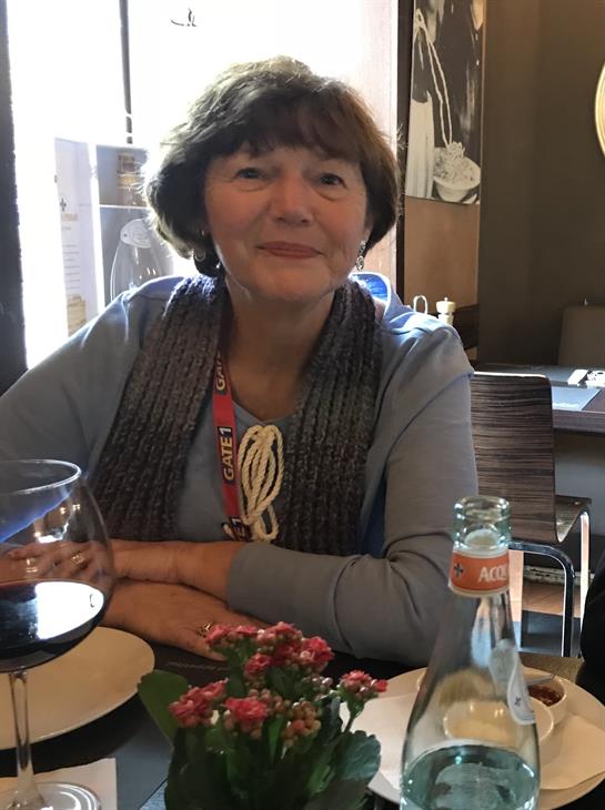 Woman sits at a table in a restaurant, smiling with a glass of wine and a floral centerpiece.