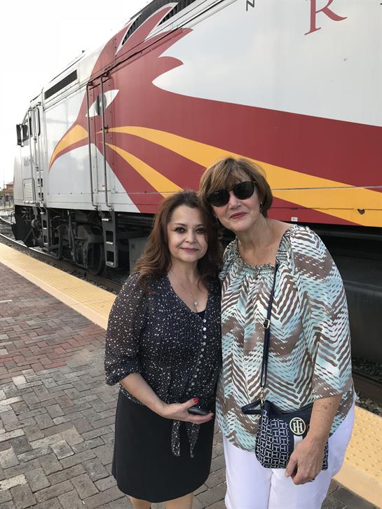 Women pose smiling at the train station while a colorful train awaits nearby.