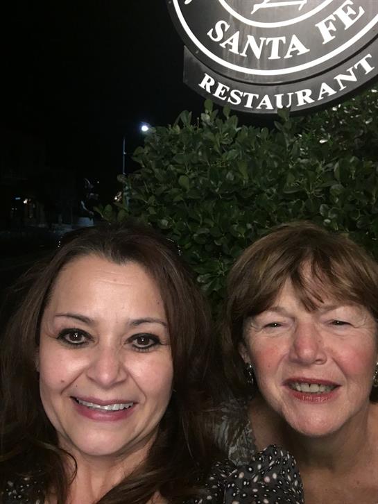 Two women pose happily in front of a restaurant sign under evening lights.