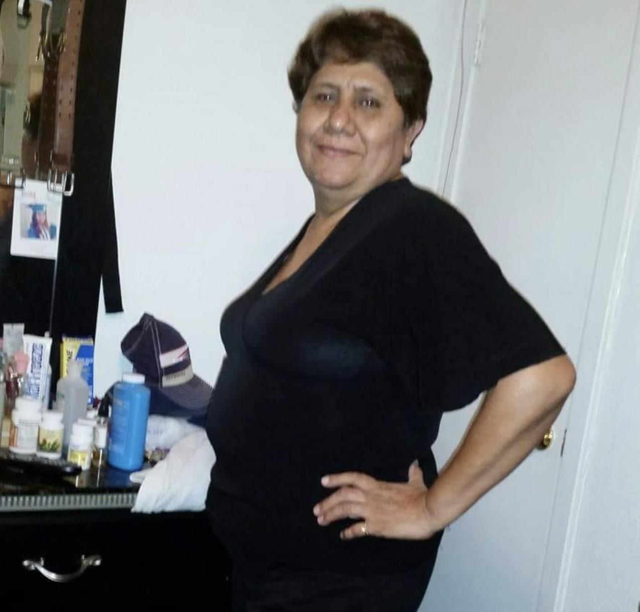 A woman stands proudly in a black outfit, showcasing her confident demeanor in a cozy room.