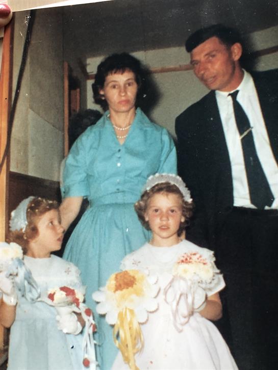 Two children in elegant dresses hold flowers as adults watch in a classic indoor setting.