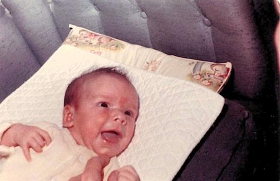 A cheerful baby is lying on a decorative pillow, smiling wide, in a warm indoor ambiance.