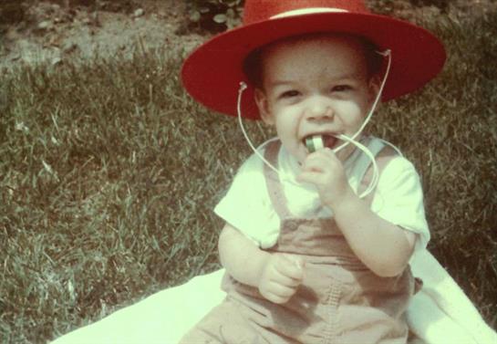 A toddler with a cheerful expression plays on grass, wearing a red hat and holding a toy in hand.