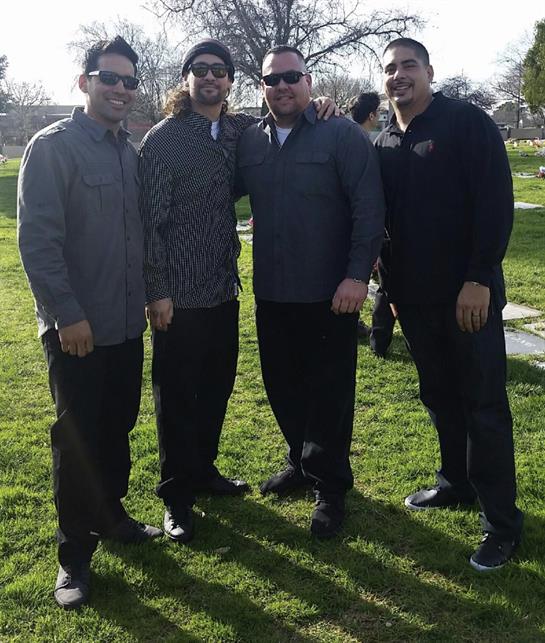 Four men stand together on green grass in a park, smiling and dressed in casual attire.